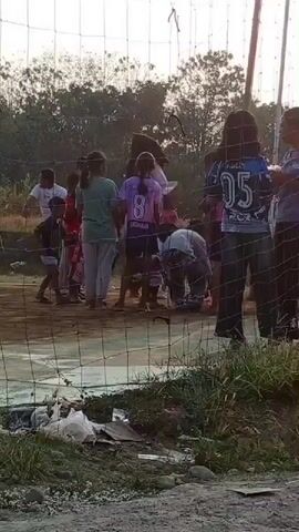A group of children and young people are gathered on a dirt court, possibly for a game or practice, with a net in the foreground. The background is lined with trees and vegetation. One child in the foreground is wearing a dark shirt and shorts, and appears to be walking or running. Another child in the middle ground is wearing a light green t-shirt and white pants. Several other children are visible in the background, some wearing athletic attire. The overall lighting suggests it is daytime, possibly late afternoon or early morning. There is some debris visible on the ground in the foreground.