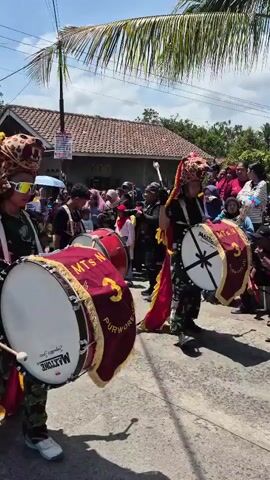 A group of people, some wearing camouflage uniforms and elaborate animal masks resembling leopards, are participating in a parade. They are playing large bass drums with red casings and white drumheads. The drums are branded with "MAXTONE." The people in the masks have decorative red and yellow fabric attached. They are walking down a paved street, with a crowd of spectators lining the sides. In the background, there are houses with tiled roofs and palm trees under a bright, sunny sky. The overall scene appears to be a lively cultural event or procession.