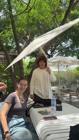 Two young adults, presumed female, are outside at a patio table under a tree. A white umbrella is positioned in front of the tree, partially obstructing the view. The person standing is holding the umbrella pole with their hands. They have dark brown hair. They are wearing a long-sleeved white shirt, black shorts, and black socks. The person is wearing a silver ring on the ring finger of their left hand. The person sitting in the chair is wearing glasses, a grey t-shirt, blue jeans, and a silver watch. They have dark brown hair and multiple necklaces. A bottle of water, two smartphones, a wallet, and a black bag are on the table. Additional tables and umbrellas are visible in the background. Another person is visible in the background. The trees are green and leafy. The camera angle is wide and stable. The person standing appears to be adjusting the umbrella to provide shade to the person sitting down.