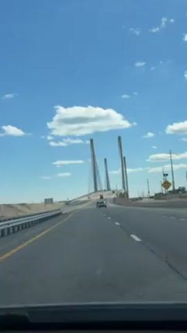 The video shows a forward-facing view from inside a vehicle driving on a highway towards a large cable-stayed bridge. The sky is blue with scattered white clouds. There are multiple cars on the road, driving in both directions. The landscape to the sides of the highway appears dry and sandy. Power lines and poles are visible on the right side of the road. The bridge has three prominent towers.
