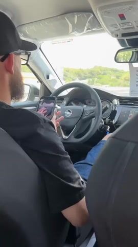 The video shows the interior of a car from a rear passenger's perspective. A man wearing a black t-shirt and a baseball cap is in the driver's seat, holding a smartphone in his left hand. The phone is mounted on the dashboard and displays a camera feed, showing the steering wheel and the driver's hand holding a vape pen. The man's right hand is on the steering wheel. The steering wheel is black with silver accents and has the Opel logo in the center. The car's dashboard displays various gauges and a digital screen. Outside the car, trees and hills are visible through the windshield. The video appears to be recorded while the car is in motion, as suggested by the slight blur of the scenery.