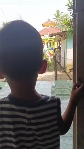 A child is looking out of a doorway at a village scene. A building that appears to be a mosque is visible in the background with a red roof and yellow walls. Flags are flying and a person is standing near the mosque. The ground is dirt and there are trees and plants. The child is wearing a striped shirt.