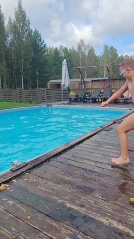 A boy jumps into a swimming pool. He is wearing blue swim trunks. The pool is outdoors and surrounded by trees and a wooden deck. There are lounge chairs and an umbrella near the pool.