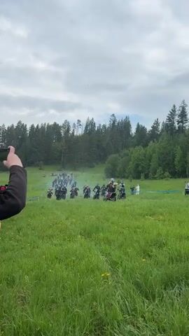 The video shows a large group of dirt bikers riding through a grassy field towards a wooded area. The bikers are wearing helmets and protective gear, and their bikes are kicking up dust and exhaust. The field is bordered by a line of trees and a few people are standing on the sidelines watching the race. Blue boundary markers are visible throughout the field. The sky is overcast and the overall environment appears to be rural.