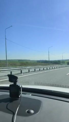 A vehicle, seen from the dashboard, is driving on a highway. The highway has guardrails on both sides. Two large trucks are visible on the highway ahead. The sky is clear and blue, with some distant clouds. The surrounding landscape is green and rural, with fields and trees. The sun is shining brightly.