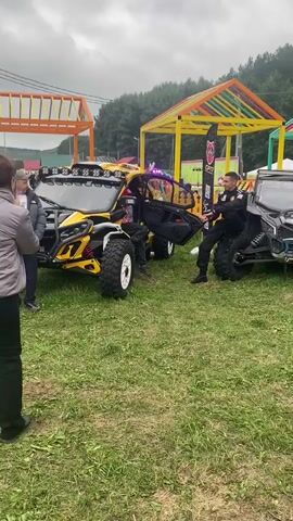 The video shows an outdoor event with several all-terrain vehicles, also known as ATVs or side-by-sides, on display. People are seen walking around and interacting with the vehicles. In the foreground, a yellow and black ATV has its door open, and two individuals, one in a dark shirt and hat, and another in a dark uniform with a badge and a hat, appear to be engaged in a conversation or demonstration near the driver's seat. To the right of this ATV is a black and blue ATV, and further to the right, a black ATV with red accents. In the background, colorful canopy structures are visible, along with trees and a forest. A person holding a camera is also visible filming the event. The ground is covered with green grass, suggesting an outdoor park or field setting. The sky appears overcast.
