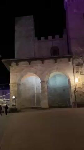 The video shows a public square at night, possibly in a European town. In the foreground is the square. A large stone building is to the left with arched passageways. A tall tower is next to the building. To the right, a large Christmas tree is brightly lit with white lights. Behind the Christmas tree is a church. The video pans right to show the church more fully. The church is made of light-colored stone and has a large round window above the doorway. There are steps leading up to the doorway. The tower is illuminated with purple light and a building behind the Christmas tree has a red light on it. A person can briefly be seen taking a photo of the Christmas tree. The general atmosphere is quiet and peaceful.