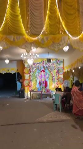 The video shows a decorated altar with a statue of the Hindu deity Ganesha. The altar is adorned with flowers and yellow and white drapes. Two large chandeliers hang from the ceiling. A group of people are seated on chairs in front of the altar, facing away from the camera. The ground is paved and appears to be outdoors.
