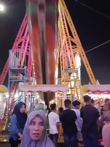 The video opens with a shot of a carnival ride, possibly a drop tower or a pendulum ride, at night. The ride structure is tall and triangular, with yellow and red accents. Below the ride, there are several stalls or booths with white canopies. A crowd of people is gathered around, some looking towards the ride and others interacting with each other. The lighting is dim, with string lights illuminating the area. As the video progresses, the camera pans across the crowd, showing various individuals, including women in hijabs and men in casual clothing. The atmosphere appears festive and busy, typical of a night market or amusement park. The camera then focuses on a group of people in the foreground, with one woman in a light-colored hijab looking towards the camera. The background is dark, indicating it's nighttime, with some trees visible in the distance. The overall scene depicts a lively gathering at a nighttime event.