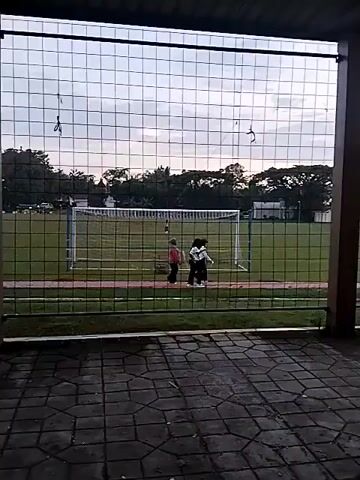 The video shows an outdoor scene, likely a sports field, viewed from behind a wire fence and a paved, covered area. Two people are walking from left to right across a green grass field in the mid-ground. A white soccer goal is visible on the left side of the field. In the background, there are trees and what appear to be buildings. Two motorcycles (scooters) are parked on the paved area in the foreground on the right. The sky suggests it is late afternoon or early evening. The overall setting appears to be a recreational or community area in Matesih, Indonesia. No visible text is present.
