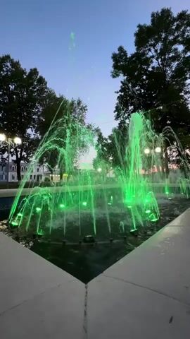 A fountain spraying water in green light is in the center of the frame. There are trees in the background with some buildings and lights visible behind them. The sky is a deep blue, indicating it is evening or twilight. The fountain has multiple jets of water, some shooting high into the air, while others arc outwards. The water is illuminated by green lights embedded in the fountain's base. The fountain is set within a paved area with a light-colored border. The overall scene is peaceful and serene, with the illuminated water creating a striking visual effect against the darkening sky.