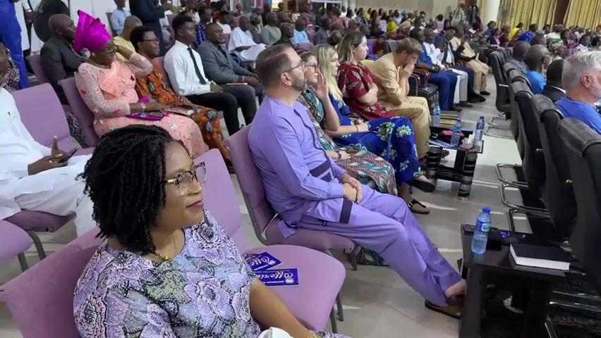 The video shows an indoor gathering of a large group of people seated in rows of chairs. The chairs are purple and arranged in sections. In the foreground, a woman with dark, braided hair and glasses is seated and looking to her right. To her right, a man in a purple matching outfit with a collarless shirt and pants is seated, also looking to his right. To his right, a woman with blonde hair and glasses is seated, looking forward. Further back, more people are visible, both men and women, dressed in a variety of clothing, some in formal wear and others in more casual attire. Many are looking towards the front of the room where a raised stage area with a red carpet is visible. On the stage, a person in a blue suit is standing and appears to be speaking. There are some decorations on the stage, including a tall white structure wrapped with red and white streamers. The overall environment appears to be a large hall or auditorium, with high ceilings and bright lighting. Several water bottles and books are visible on small tables placed between the rows of chairs. The camera pans slightly to the right, showing more of the congregation and the stage area.