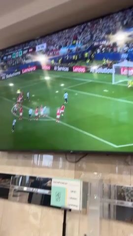 The video displays a large television screen showing a soccer match. The score is visible on the screen as 0-0. The game appears to be between Argentina and Austria, based on the jerseys and markings on the screen. In the foreground, a sign with text is partially visible. The sign has sections labeled "going", "refreshed", and "carefree". The overall environment appears to be indoors, possibly a lounge or public space with a wall of reflective material below the television. The camera is positioned at a slight upward angle, capturing the screen and a portion of the room. The footage pans across the screen showing different players and moments of the game. One notable close-up is of player Lionel Messi. The scene is well-lit, allowing for clear visibility of the television content.