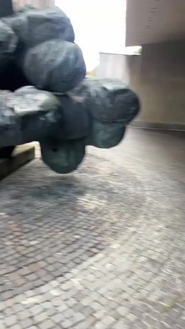 This video shows a bronze sculpture with multiple figures and hands. The sculpture is located in Banská Bystrica, Slovakia, in front of a building with large windows. The scene is outdoors, with cobblestone paving. In the background, there is a memorial flame burning in a brazier. The sculpture appears to be a memorial, possibly to victims of war or persecution, given the somber tone and the presence of the flame. One section of the sculpture depicts a person embracing another, while other parts show many outstretched hands reaching out. The lighting suggests it is daytime, but the weather is overcast. There is no visible text in the video.