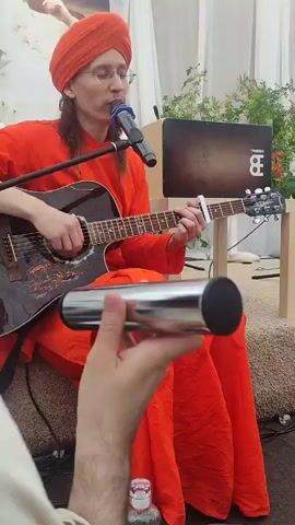 A person wearing an orange turban and robe plays an acoustic guitar while singing into a microphone. They are seated on the floor. In the foreground, a hand holds a cylindrical percussion instrument. In the background, another person stands and faces away from the camera, and a wooden box with a logo rests on a table. The environment appears to be outdoors or in a tent-like structure with white fabric draping.