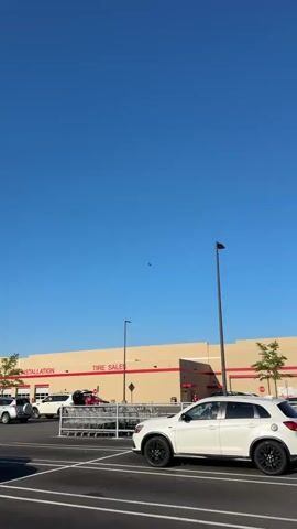 The video shows an outdoor scene during the day. The sky is clear and blue. A commercial airplane is visible high in the sky. Below, there's a Costco building with signage that reads "Installation" and "Tire Sales". A parking lot is in front of the building with parked cars, including a white SUV. Shopping carts are lined up. People can be seen near the carts. A street light is also visible.