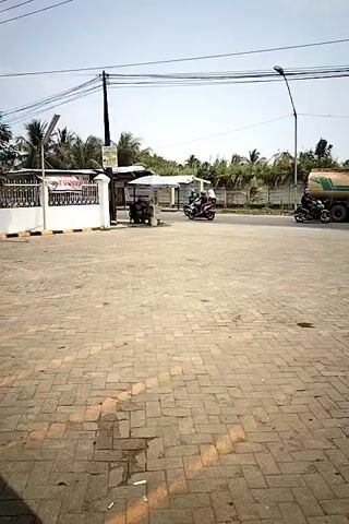 The video shows a paved area with brick paving stones. In the background, there is a street with a few vehicles. On the left side of the image, there is a white fence and a small building with a canopy. Under the canopy, there appears to be a small food stall or cart with people seated around it. Two motorcycles are visible driving on the street, one passing by the stall and the other further down the road. A white car is also seen on the street. Palm trees and other foliage are present in the background, along with a concrete wall. Overhead, there are electrical wires and a street lamp. The sky is clear and bright, indicating daytime.