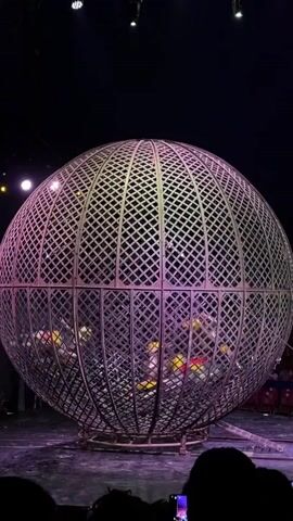 A motorcycle is being driven inside a large metal sphere. The sphere is on a stage with purple lighting. There are people in the audience watching. The motorcycle is moving around the inside of the sphere. The motorcycle is yellow and black. The sphere is made of metal bars and is open in places.