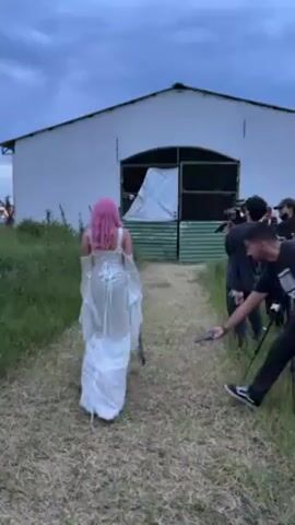 A video shows a person dressed in a white, ornate dress and pink hair walking away from the camera towards a white barn. A group of people are next to the path, setting up camera equipment, presumably to film the person. One man crouches and appears to be examining the ground in front of the person walking. The sky is overcast.