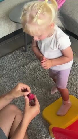 A person's hands are holding a small red object, possibly a strawberry, and are offering it to a young child. The child, a blonde girl with braided hair and wearing a white t-shirt and purple shorts, is standing on a yellow platform. She is looking down at the object, her hands clasped in front of her. The scene appears to be indoors, with a gray rug and furniture visible in the background.