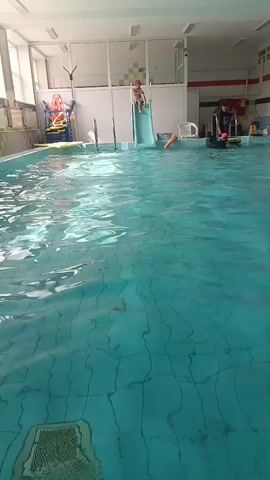 A child slides down a blue slide into a swimming pool. Another child, wearing a pink swim cap, swims in the pool. An adult stands near the edge of the pool, facing away from the camera. There are various pool accessories visible, including kickboards and a raised platform. The room appears to be a public indoor swimming pool.