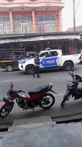A police officer directs traffic on a street. A white police vehicle with blue and white markings is parked behind him. A white SUV is driving down the street. Several motorcycles are parked on the side of the road. Buildings are visible in the background.