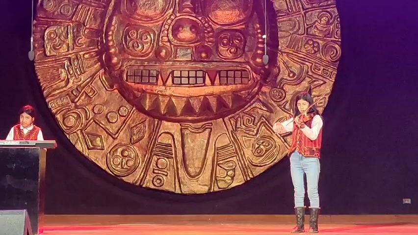 Two young people are performing on a stage. One is playing a violin, while the other is seated at a keyboard. Both are wearing similar vests. Behind them is a large, round, golden prop, possibly representing a sun or other ancient symbol. The stage has a dark backdrop and a wooden floor. The scene is well-lit.