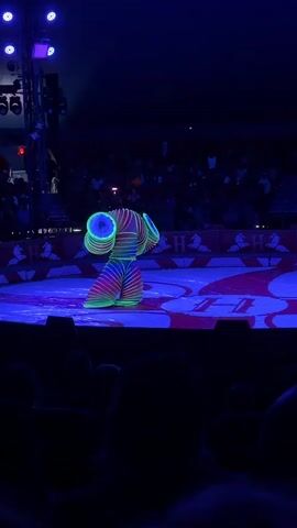 A performer in a brightly colored, neon slinky-like costume is doing a flexible dance routine on a circular stage under blue lights. The performer's costume is illuminated by blacklights, making the neon colors glow. The performer's body is contorted into various positions, including standing upright, bending over, and twisting. The stage is decorated with a red and white checkered pattern, and there are red and white striped barriers around the edge of the stage. In the background, a dark audience is visible, seated in bleachers. The overall atmosphere is that of a circus or a performance show.