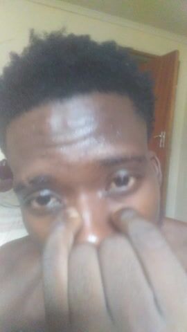 The video features a close-up, selfie-style recording of a young man with dark skin and short, dark, curly hair. He appears to be shirtless or wearing a very light-colored garment that blends with the background. The video is shot from a slightly low angle, looking up at his face.

At the beginning (0:00-0:01), he is smiling widely, showing his white teeth and a bit of his tongue. He then shifts his expression (0:01-0:02) to a more relaxed smile, still showing his teeth, and then briefly looks off-camera before returning his gaze. Around 0:02-0:03, he closes his mouth slightly, maintaining a pleasant expression, and turns his head slightly to his left, revealing the side of his face and a hint of a short beard or stubble along his jawline. His skin appears smooth and somewhat shiny.

The background is simple, consisting mainly of a light, muted color (possibly a wall or bedding), with a darker brown object, which appears to be a wooden door or furniture, visible behind his right shoulder towards the end of the clip. The lighting is bright and even. The man speaks briefly at the very beginning of the clip, saying "Fresh cut," suggesting he is showing off a recent haircut.