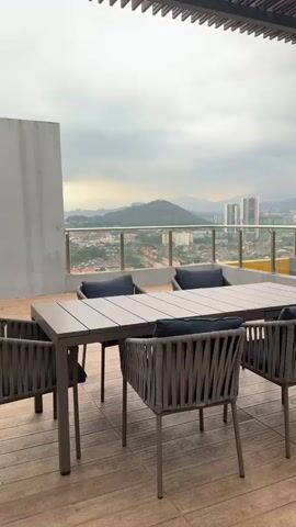 This is a vertical, medium shot of an outdoor dining set on a balcony, with a city view and mountains in the background. The dining set consists of a rectangular table and six chairs. The table is made of grey material and has a slatted surface. The chairs are made of a woven grey material with blue cushions. The balcony has a wooden floor and a glass railing. Beyond the railing, the view includes a cityscape with several tall buildings, houses, and lush green mountains in the distance. The sky is overcast and cloudy.