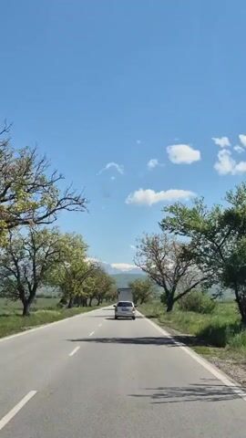 The video shows a car driving on a paved road on a clear day.  The road has yellow dashed lines down the center and solid white lines on the right shoulder.  Trees line both sides of the road, and there is a green field on either side of the trees.  A white car is driving ahead on the same road. In the distance, mountains can be seen.  The sky is blue with a few small white clouds.