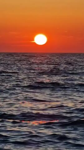 A vertical video shows the sun setting over the ocean. The sun is a bright white disc against an orange sky. The ocean is dark blue with small waves. The sunlight reflects on the water in a shimmering path.