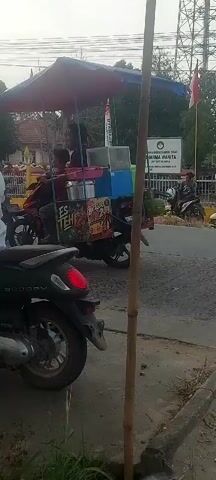 The video begins with a close-up of a motorcycle parked on the side of a road. In the background, a sign reads "DHARMA WANITA". Several people on motorcycles are visible, some wearing red shirts. One person is wearing a conical hat and a red shirt, riding a motorcycle. Another person on a motorcycle is wearing a blue shirt. The road appears to be wet, suggesting recent rain. The overall scene depicts a procession or gathering of people on motorcycles, possibly a parade or event. The weather appears to be overcast.
