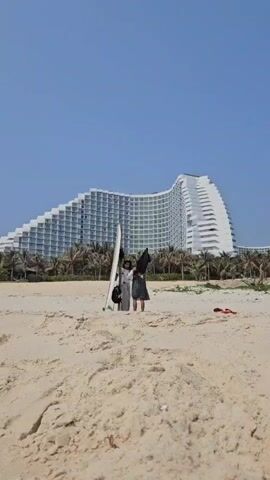 A bright, sunny day at the beach features two people standing beside a white surfboard. The people appear to be interacting, possibly adjusting their belongings. They are standing on the sandy beach, with a large, uniquely designed white hotel in the background, lined with palm trees. A pair of red sandals sits on the sand.