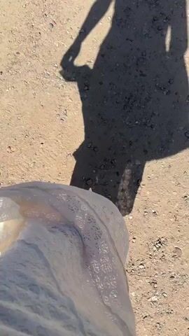 The video shows a first-person perspective of a person walking on a dirt and gravel path on a sunny day. The person is wearing a long, white garment and Adidas shoes. The shadow of the person in the video is cast onto the path, revealing that they are holding an object in their hand, possibly a phone or camera. The ground is covered with small rocks and pebbles. There is some faint ambient noise in the background.