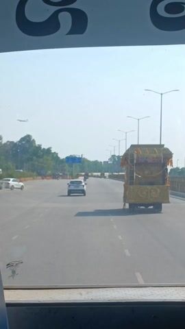 The video, shot from the interior of a bus, captures a scene on a wide, multi-lane highway during daylight. The bus, possibly an older model, is actively moving, as evidenced by the changing scenery. In the foreground, the bus's interior is visible, including the dashboard, steering wheel, and some passenger items. A man's hand is partially visible on the steering wheel, suggesting he is driving the bus. A backpack is resting on the seat next to the driver's area. Through the windshield, the view unfolds. Several other vehicles are seen on the road, including a white SUV and a small car. A noteworthy vehicle, elaborately decorated with what appears to be religious or cultural symbols, is traveling in the same direction as the bus. Streetlights line the sides of the highway, and green foliage is visible in the background, adding to the sense of an open road. The overall atmosphere is bright and sunny.