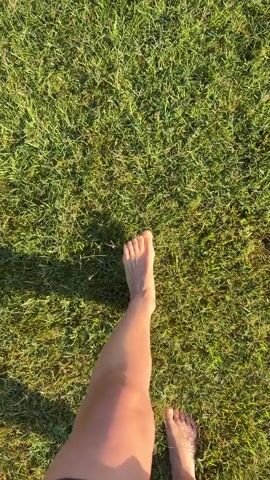 The video shows a first-person perspective of someone walking barefoot on green grass. The sun casts long shadows, indicating it might be late afternoon or early morning. The focus is on the person's right leg and foot as they move forward through the grass. The environment is a grassy outdoor area, likely a lawn or park. No other people or objects are clearly visible.