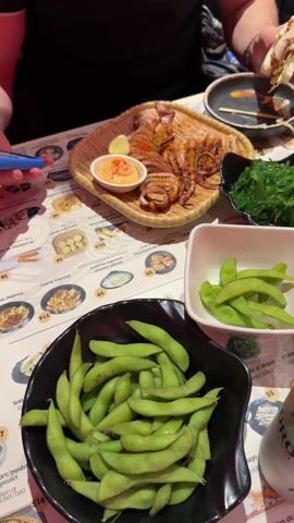 The video shows a table laden with Japanese food and drinks. There's a dish of grilled squid with a side of sauce, a bowl of seaweed salad, two bowls of edamame, and a plate of gyoza. Two cans of Sapporo Premium beer are also on the table. Two people are visible, one holding a sandwich-like object and the other holding a phone and focusing on the food. The table is printed with a menu. The environment appears to be a restaurant.