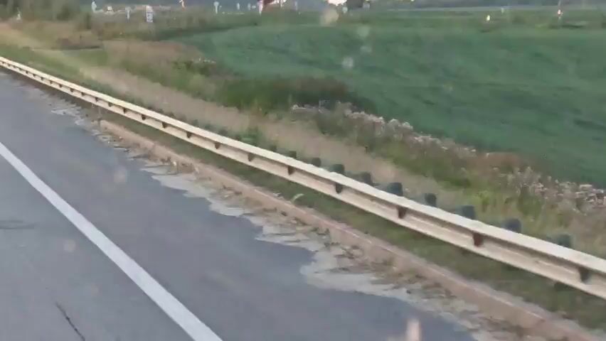 The video shows a hot air balloon in the sky at sunset. The balloon is red and white. It is in a field with trees in the background. A road is visible in the foreground. The balloon appears to be ascending.