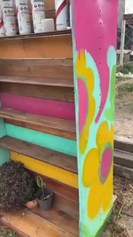 The video shows a man spray painting. The video begins with a close-up of a colorful shelf and then pans out. The shelf has a light blue frame, with wooden shelves colored orange, pink, light blue, and yellow. There are several cans of spray paint on the top shelf, and a couple of plants on the bottom. Beside the shelf is a brightly colored door with pink, yellow, green, and white paint. A man in a white t-shirt and khaki pants stands in front of the door with a spray paint can in his hand. In the background, there is a white pickup truck and a building with a closed gray garage door. The man sprays paint on the door, then walks toward the camera.