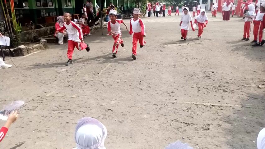 The video shows a group of children participating in an outdoor activity, likely a game or competition, in Sukaraja, Indonesia. The children are dressed in matching red and white uniforms, suggesting they are part of a school or organized group. The scene appears to be a dirt or gravel area, possibly a schoolyard or a public space.

Several children are actively engaged in a task involving what appear to be small bottles placed on the ground. Some children are bending down to interact with these bottles, while others are in motion, possibly running or moving between stations. One child in the foreground is wearing a headscarf, indicating potential cultural context. The overall atmosphere seems energetic and focused.

The time of day appears to be daytime, with sunlight casting shadows on the ground, suggesting clear or partly cloudy weather. The setting is rural or semi-rural, consistent with the location context of Sukaraja, Indonesia.

Visible text on the uniforms includes "KARANJA" and "TEN BOGOR," likely indicating the name of a school or organization and its location. The children's expressions are difficult to discern clearly, but the activity suggests a sense of competition and engagement.