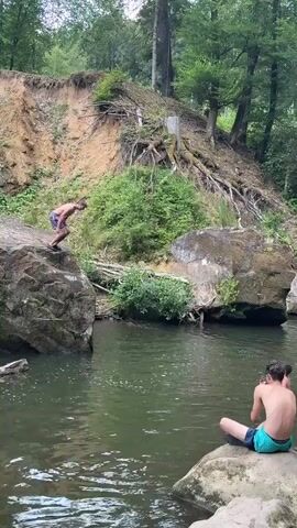 At timestamp 00:00, a person in dark-colored shorts is mid-air, having jumped from an elevated, dirt embankment with exposed tree roots into a body of water. The water is green and appears to be a river or stream, with large rocks on either side. Another person is seated on a rock in the foreground, facing away from the viewer, wearing light blue and green patterned shorts. In the background, more trees and vegetation are visible, with the embankment continuing to the left.

At timestamp 00:01, the person who jumped is now impacting the water, creating a large splash. The seated person in the foreground remains in the same position.

At timestamp 00:02, the splash continues to expand, and the person is now submerged beneath the water.

At timestamp 00:03, the person has surfaced and is swimming in the water.

At timestamp 00:04, the person is still in the water, appearing to be treading water or swimming. The seated person and the environment remain unchanged.