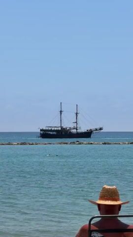 The video shows a man in a straw hat sitting in a chair, facing the ocean. In the background, there is a pirate ship with two masts. The ocean is calm and blue, and the sky is clear with only a few clouds. There are a few people swimming in the ocean. The ship is located behind a rock jetty. The man is not moving, just observing the scene.