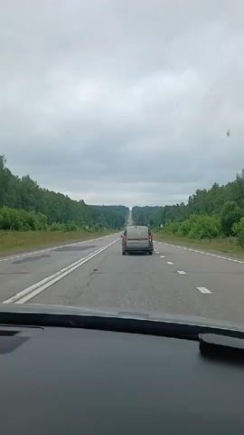 A first-person view from inside a vehicle shows a grey van driving on a two-lane road. The road is surrounded by green trees on both sides, and the sky is cloudy. The van is in the right lane, and the viewpoint vehicle is in the left lane. The road appears to be somewhat bumpy, with some visible patches and cracks. There are traffic signs on the side of the road, but they are too distant to be read. The video is short and appears to be a recording of a car trip.