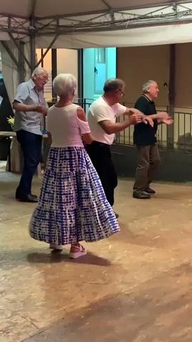 This video shows four older adults dancing outdoors under a canopy. Two men are in the background, one on the left wearing a light blue shirt and dark pants, facing a woman. Another man in the background on the right wears a black shirt and khaki pants. In the foreground, a woman with short blonde hair wears a white off-the-shoulder top and a long blue and white patterned skirt. She is dancing with a man in a white t-shirt and black pants. The floor is a light-colored, possibly wooden surface. The lighting suggests it is evening. The overall scene depicts a social gathering with dancing.