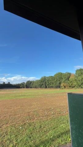 The video shows a view of a golf driving range on a sunny day. The sky is blue with a few scattered clouds. In the foreground, the edge of a roof or overhang is visible, casting a shadow. The driving range itself is a large, open field with patches of brown and green grass. Numerous white golf balls are scattered across the field, and there are several small orange flags marking targets. A line of trees borders the far end of the driving range, with some trees showing autumnal colors. The overall scene depicts a clear and pleasant outdoor environment suitable for practicing golf.