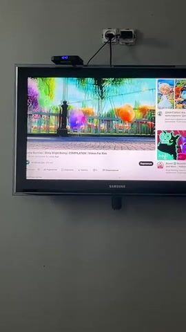 The video shows a television screen displaying a YouTube video. The video features several animated, colorful rabbit-like characters in an outdoor setting with a fence and trees in the background. The characters are interacting, with some of them appearing to bounce or move around. The YouTube interface is visible on the bottom portion of the screen, indicating the video title is "Sunny Bunnies | Shiny Bright Bunny | COMPILATION | Videos For Kids." There are also suggested videos displayed on the right side of the screen. The television itself is a Samsung brand and is mounted on a wall. Above the television, there is a cable box and two electrical outlets.