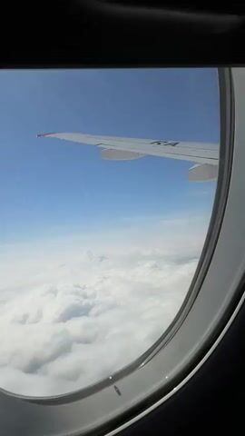 This is a POV shot from an airplane window, looking out at the sky and clouds. The wing of the airplane is visible in the upper right corner, extending into the frame. Below the wing, a vast expanse of white, fluffy clouds stretches out to the horizon. The sky above is a clear, bright blue. The camera is pointed downwards and slightly forward, capturing the vastness of the sky and the layer of clouds below. The overall impression is one of flight and a serene view from above the clouds.