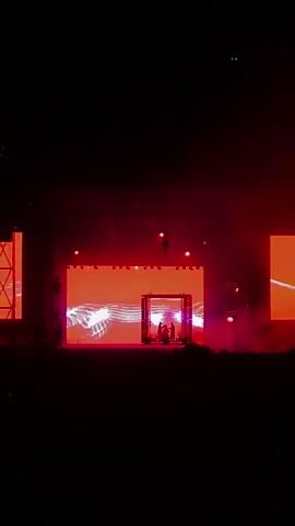 A concert stage is illuminated by purple and pink lights. Two figures stand in the center of the stage, silhouetted against a large screen displaying abstract visuals. Several red lights are suspended above the stage, and smoke drifts across the scene. The audience is barely visible in the darkness at the bottom of the frame. The video then cuts to a dark scene with red lights and smoke. Finally, the lights change to red and the text "ESTRELLA DAMM" appears on the screen, with the audience visible in silhouette.
