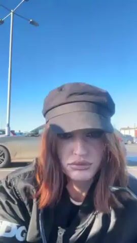 A person with shoulder-length auburn hair and wearing a black cap, black jacket, and black track pants with white stripes is the focus of the video. They are standing outdoors in a parking lot with several vehicles visible in the background. A tall streetlight is positioned on the left side of the frame. The person is holding a clear bag with items inside. Initially, they are close to the camera, then move away, twirling and dancing while holding the bag. The sky is clear and blue.